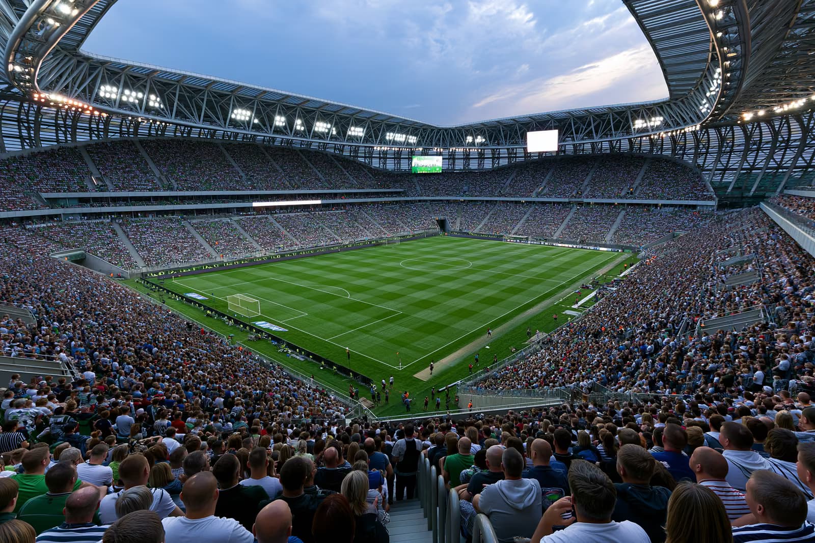 Velký fotbalový stadion plný fanoušků pod reflektory za letního večera.