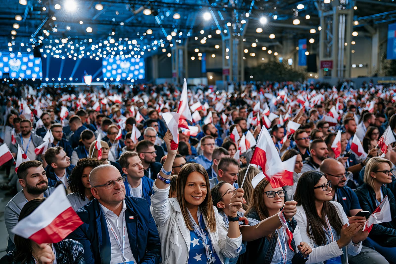 Zaplněný dav v výstavní síni mávající malými vlajkami pod ostrým osvětlením při zahájení kampaně AfD v Magdeburgu, v dálce pódium.